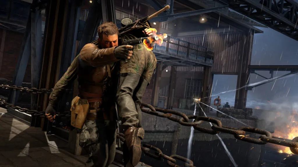 In a scene from Sniper Elite: Resistance, Harry Hawker wields a silenced pistol, whilst carrying another soldier over his shoulder on a rainy dockyard. He is walking over a chained bridge with gunfire zipping all around him.